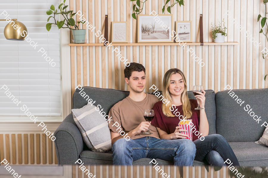 A young couple sitting on the sofa, watching a movie, and enjoying red wine and popcorn stock photo with image ID: 3eb9684d-4412-4745-ad9e-d2a75eda96ee