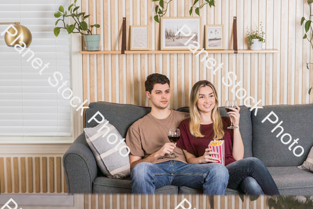 A young couple sitting on the sofa, watching a movie, and enjoying red wine and popcorn stock photo with image ID: 3eb9684d-4412-4745-ad9e-d2a75eda96ee